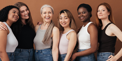 Multiple women, all of different ages and ethnicities, posing and smiling together in tank tops and jeans.