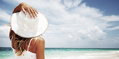 A woman grasping her wide brim white hat on the beach as she looks at the clouds.